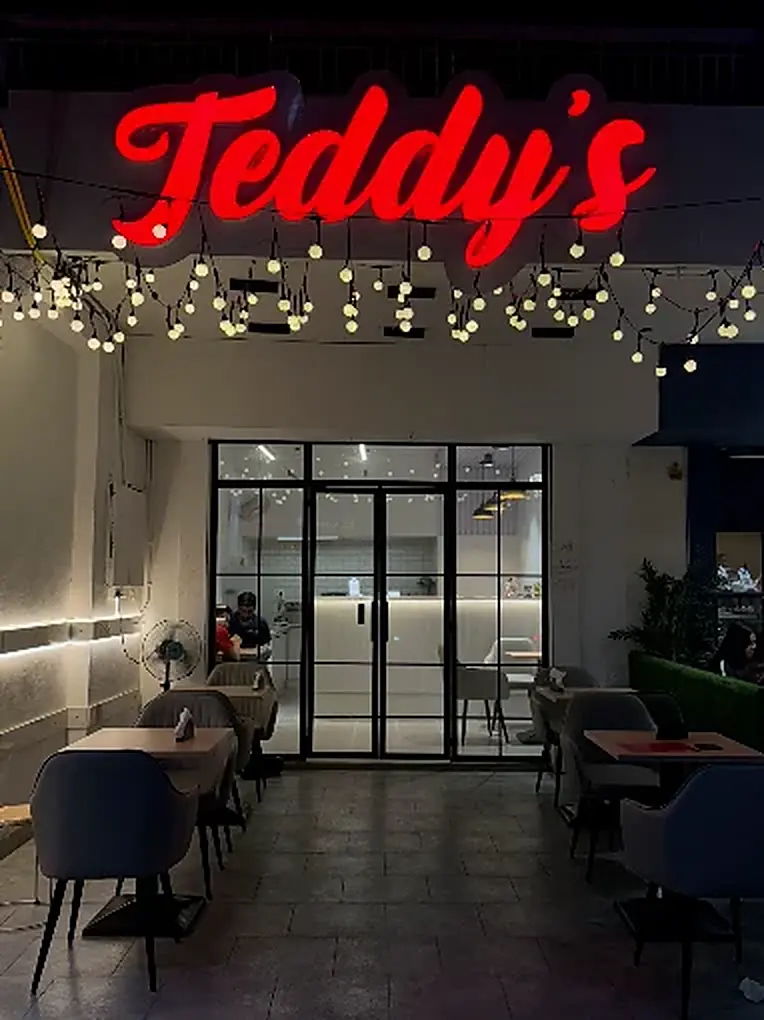 Teddy's from the road at night — the neon sign, strings of bulb lights over empty tables, and the lit glass front behind