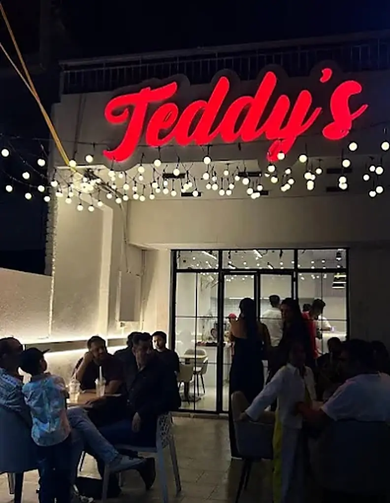 The Teddy's shopfront at night — the red neon sign above strings of bulb lights, with people at tables outside